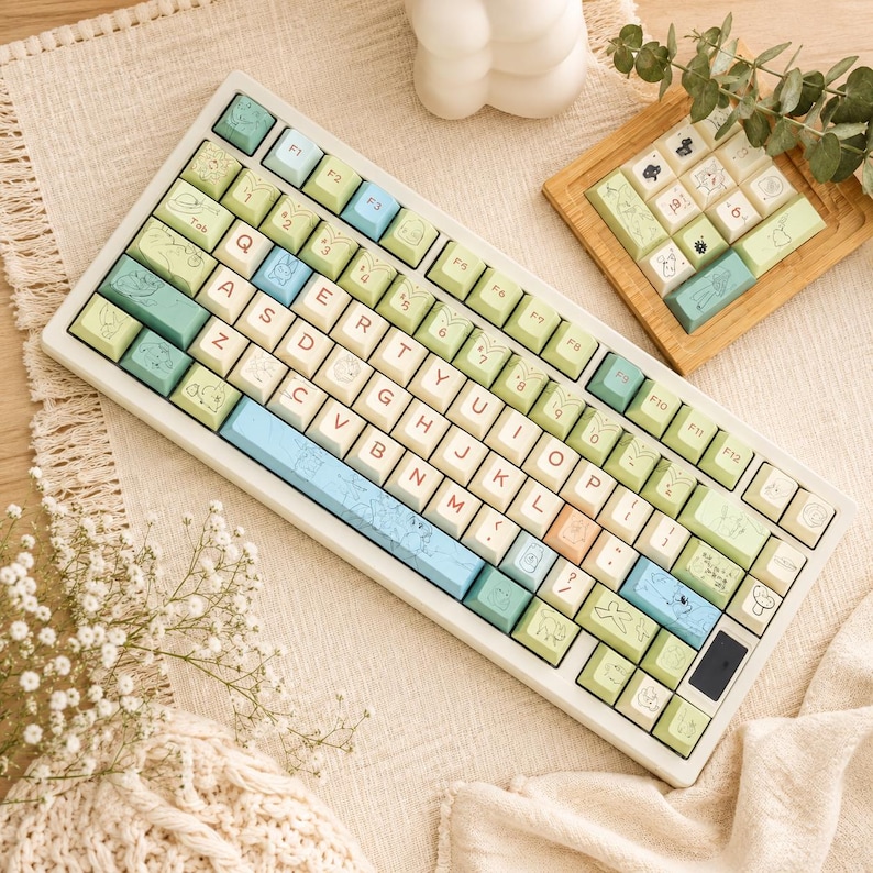 May include: A mechanical keyboard featuring a white frame and a custom keycap set in shades of sage green, light blue, and cream. The keys display QWERTY layout letters in red, along with various small line-art illustrations of woodland creatures.
