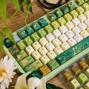 Könnte beinhalten: Eine mechanische Tastatur in Grün und Creme mit individuell gestalteten Tastenkappen, die botanische Illustrationen von Lotusblumen, Blättern und Kranichen zeigen. Die Tasten tragen eine Standard-QWERTY-Beschriftung.