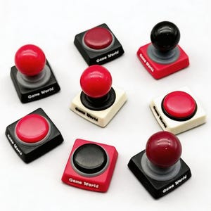 Puede incluir: Una colección de mini mandos de juego estilo joystick. Los mandos presentan una variedad de colores, incluyendo rojo, negro y blanco. Cada mando tiene una parte superior redondeada y una base cuadrada. El texto "Game World" está impreso en la base de algunos mandos.