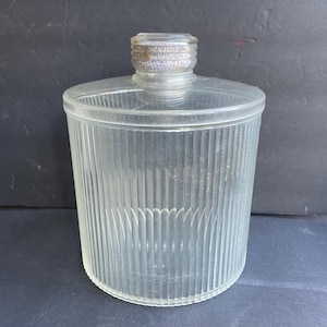 May include: A clear, cylindrical glass container with vertical ribbed detailing. The container has a flat, circular lid and a threaded metal top. The glass is transparent, allowing a view of the contents. The design is simple and functional.
