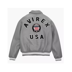 May include: A gray bomber jacket with white and gray striped cuffs and hem. The back features the text "AVIREX" above a circular emblem and "USA" below. The emblem includes a red, white, and blue design.