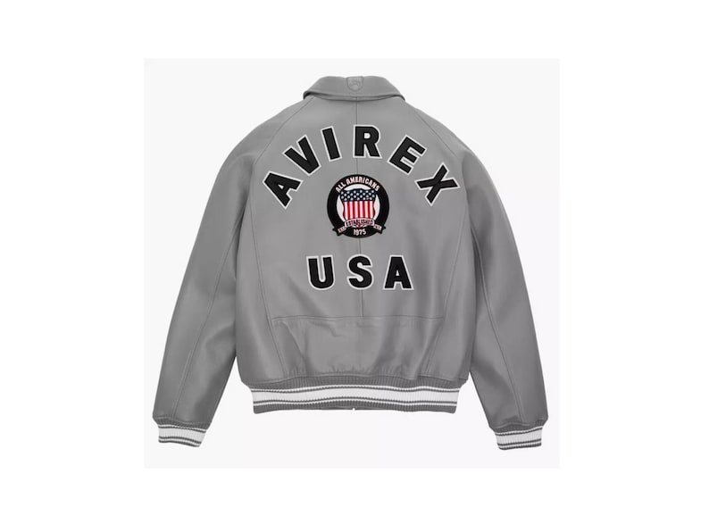 May include: A gray bomber jacket with white and gray striped cuffs and hem. The back features the text "AVIREX" above a circular emblem and "USA" below. The emblem includes a red, white, and blue design.
