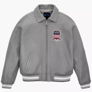 May include: A gray leather jacket with a zip-up front, collar, and ribbed cuffs and hem. The jacket features a patch with red, white, and blue details. The brand name "MAD" is visible on the collar.