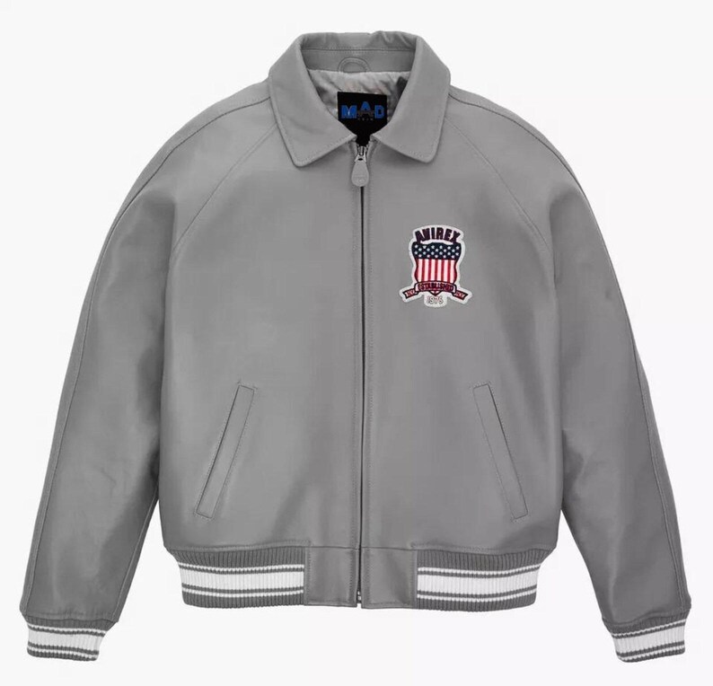 May include: A gray leather jacket with a zip-up front, collar, and ribbed cuffs and hem. The jacket features a patch with red, white, and blue details. The brand name "MAD" is visible on the collar.