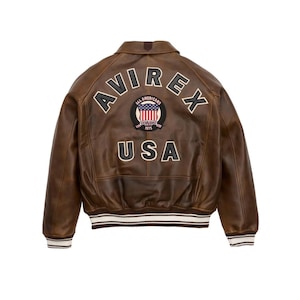 Pu&ograve; includere: Giacca in pelle marrone con la scritta "AVIREX" ad arco sulle spalle e "USA" sotto. Una toppa circolare con la bandiera americana &egrave; centrata tra le parole. La giacca ha una fascia a righe bianche e marroni in vita e sui polsini.