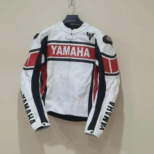 Puede incluir: Chaqueta de moto blanca y negra con detalles rojos. La chaqueta lleva la palabra "YAMAHA" en rojo en el pecho y en las mangas. Cuelga de una percha negra.