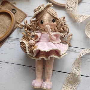 May include: A handmade crochet doll with a pink dress and a matching hat. The doll has curly brown hair and is holding a small basket. The doll's dress is trimmed with a lighter pink color. The doll is wearing pink shoes.