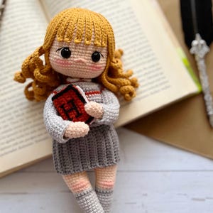 Könnte beinhalten: Eine Häkelpuppe mit lockigem goldenem Haar, bekleidet mit einem grauen Pullover und Rock mit rotem Streifen, hält ein kleines rotes Buch mit dem Buchstaben H auf dem Einband. Die Puppe hat schwarze Augen, rosa Wangen und helle Haut.