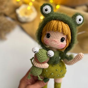 May include: A handmade crochet doll wearing a green frog hat and dress, holding a small green frog. The doll has blonde hair, rosy cheeks, and large black eyes. The frog doll has white eyes and a red collar.