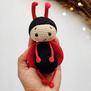 May include: A handmade crochet ladybug doll with a black body, red wings, and a beige head. The doll has black eyes, rosy cheeks, and red antennae. The doll is held in a hand.