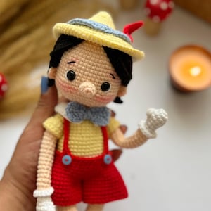 May include: A handmade crochet doll of Pinocchio, featuring a yellow hat with blue trim, black hair, and a red feather. The doll wears a yellow shirt, red shorts with blue buttons, and a blue bow tie. White gloves complete the look.