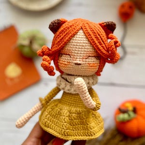 Puede incluir: Muñeca de ganchillo hecha a mano con cabello naranja y un vestido amarillo. La muñeca tiene una bufanda color crema y sostiene un objeto pequeño y de color claro. La muñeca tiene los ojos cerrados y una expresión serena. El fondo incluye una pequeña calabaza naranja y un bloc de notas.
