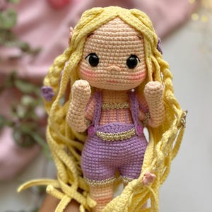 May include: A handmade crochet doll with long, flowing yellow hair and a sweet expression. The doll wears a lavender outfit with gold accents. The doll's arms are raised, and it has a light pink blush on its cheeks. The doll is a charming handmade item.