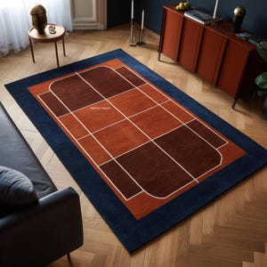 Puede incluir: Alfombra rectangular con borde azul marino, que enmarca un diseño de formas geométricas en tonos marrón y naranja. Líneas blancas definen las formas. La alfombra está colocada sobre un suelo de madera.