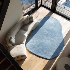 Könnte beinhalten: Ein geschwungenes, cremeweißes Sofa mit drei passenden Zierkissen neben einem großen, unregelmäßig geformten, hellblauen Teppich. Die Möbel stehen auf einem hellen Holzboden in einem Raum mit großen Fenstern und natürlichem Lichteinfall.