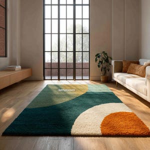 May include: A rectangular rug with an abstract design in a living room. The rug has a teal, yellow, white, and orange colour scheme with geometric shapes. It is placed on a wooden floor. The text "SabhyCarpets" is visible on the rug.
