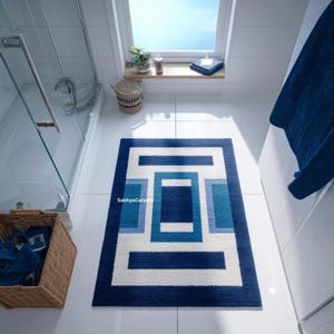 May include: A rectangular bath mat with a geometric design in shades of blue and white. The mat features a dark blue border, with a central pattern of rectangles and squares in varying shades of blue and white. The text "Sabhyacarpets" is visible.