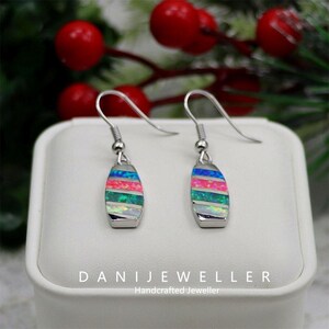 May include: A pair of silver dangle earrings with a colorful, striped design. Each earring features a rectangular, opal-like stone with horizontal bands of blue, pink, green, and white. The earrings are displayed on a white surface with text that reads "DANIJEWELLER Handcrafted Jeweller."