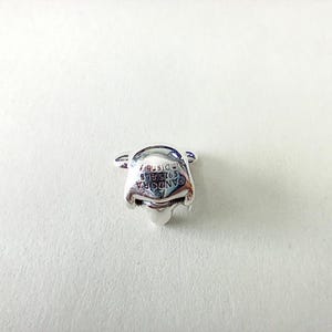 Może przedstawiać: Srebrny charms w kształcie twarzy Kubusia Puchatka. Charms ma wygrawerowany napis "PANDORA S925 ALE &copy;Disney" z przodu. Charms znajduje się na białym tle.