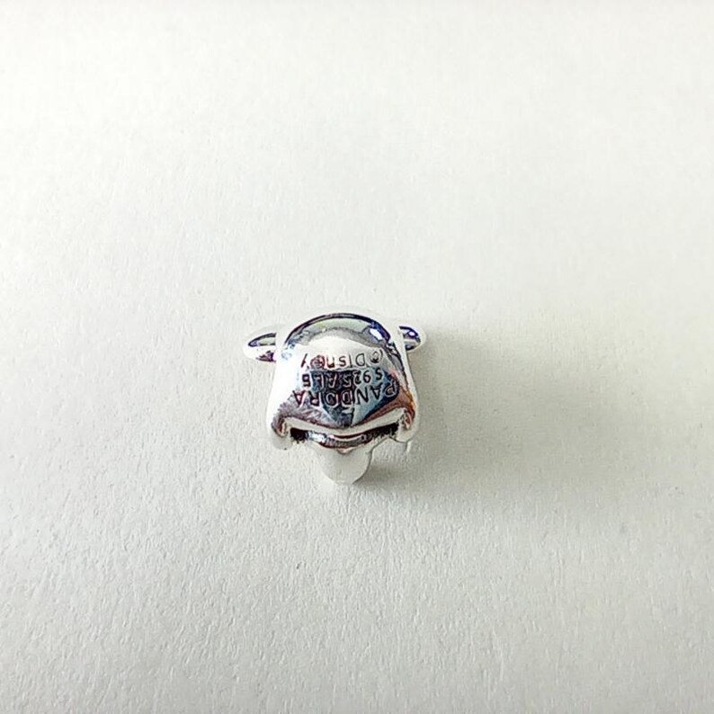 Może przedstawiać: Srebrny charms w kształcie twarzy Kubusia Puchatka. Charms ma wygrawerowany napis "PANDORA S925 ALE &copy;Disney" z przodu. Charms znajduje się na białym tle.
