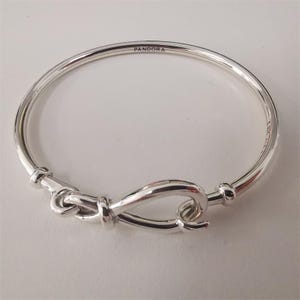 May include: A silver bangle bracelet with a knot design clasp. The bracelet has a smooth, polished finish and the word "PANDORA" is engraved on the top. The clasp is a unique, eye-catching feature.