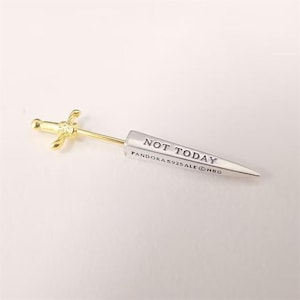 Puede incluir: Un pendiente plateado y dorado en forma de daga. El pendiente presenta una empuñadura dorada y una hoja plateada con el texto "NOT TODAY" y "PANDORA S925 ALE © HBG" grabado. El pendiente está sobre un fondo blanco.