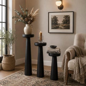 Pode incluir: Três mesas de pedestal pretas de alturas variadas dispostas em fila. A mais alta sustenta um vaso de cerâmica com flores secas, a do meio um castiçal de vidro com uma vela e a mais baixa uma pilha de três livros.