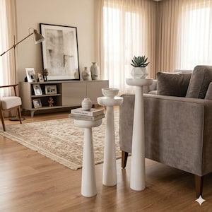 May include: Three white pedestal tables of varying heights, each with a round top. One table holds a small succulent in a geometric pot. The tables are in a living room with a neutral color scheme, a rug, and a sofa.
