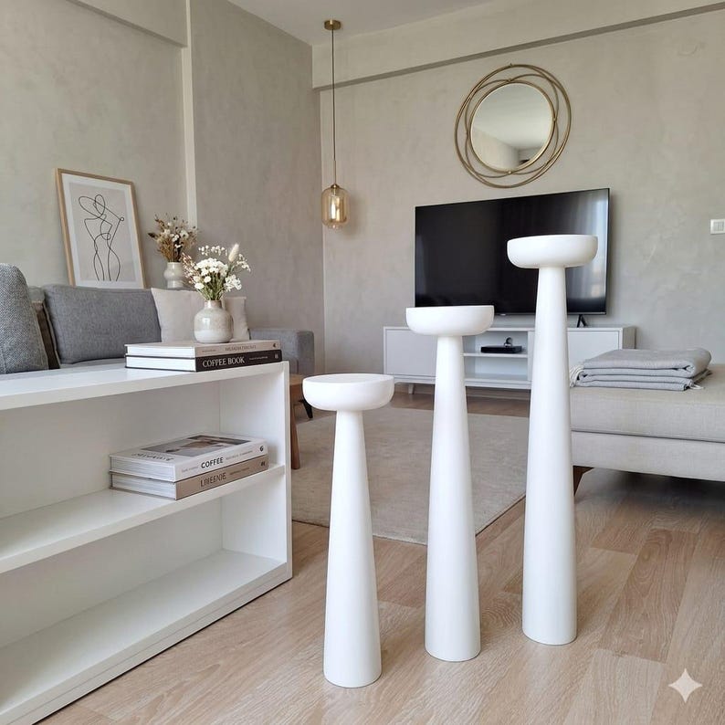 May include: Three white pedestal stands of varying heights. Each stand has a round top and a conical base. The stands are arranged in a living room setting, suggesting a decorative or display purpose. The room has a neutral color palette.