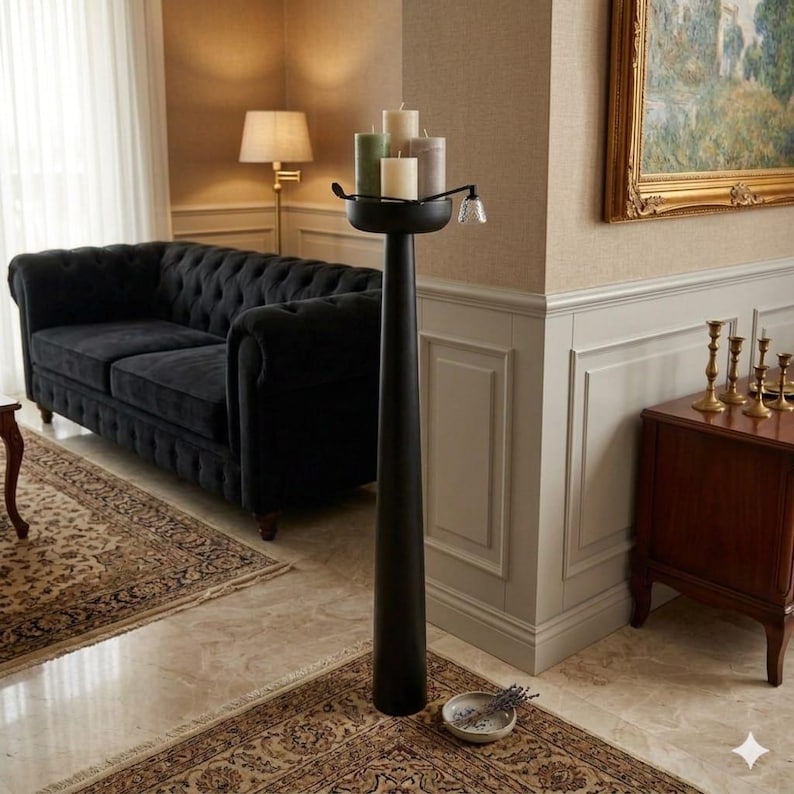May include: A tall, black metal candle holder with a round top holding several candles in shades of green and cream. The holder stands in a living room with a black sofa, patterned rug, and a painting on the wall. A small dish with lavender sits at the base.