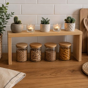 Könnte beinhalten: Ein Küchenregal aus hellem Holz trägt zwei kleine Kakteen in Töpfen, zwei Teelichthalter aus Glas mit brennenden Kerzen und eine kleine Sukkulente. Darunter stehen vier durchsichtige Gläser mit Bambusdeckeln, die verschiedene trockene Lebensmittel enthalten.