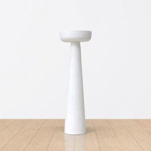 May include: A tall, white, minimalist pedestal with a round top and a conical base. The pedestal is made of a solid, opaque material and stands on a light-colored wooden floor against a white wall.