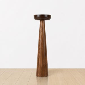 May include: A tall, wooden pedestal with a dark brown, bowl-shaped top. The pedestal has a conical shape, widening towards the base. The wood grain is visible, showcasing a natural, warm brown color. The design is minimalist and modern.