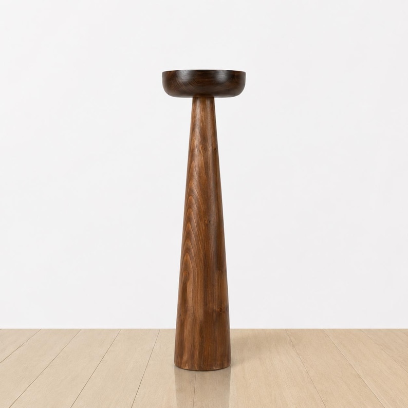 May include: A tall, wooden pedestal with a dark brown, bowl-shaped top. The pedestal has a conical shape, widening towards the base. The wood grain is visible, showcasing a natural, warm brown color. The design is minimalist and modern.