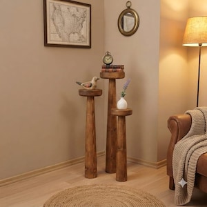 May include: Three cylindrical wooden plant stands of varying heights, each featuring a circular top and a tapered base. The tallest stand holds a small clock and books, the medium stand holds a bird figurine, and the shortest holds a white vase.