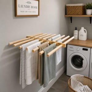 Peut inclure: Un séchoir mural en bois doté de plusieurs tiges horizontales s'étendant vers l'extérieur. Plusieurs serviettes dans des tons gris, blanc et beige sont suspendues sur les tiges. Un panneau encadré indique LAUNDRY ROOM WASH DRY FOLD REPEAT.