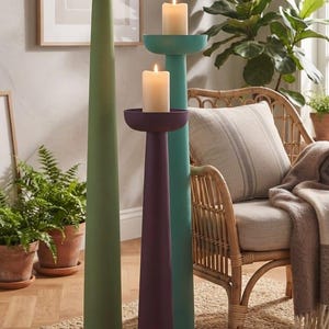 Pode incluir: Três castiçais de chão altos e esguios em verde-oliva mate, azul-petróleo e roxo escuro. Cada suporte apresenta uma plataforma superior circular larga que sustenta uma vela de pilar cor de creme. O texto "DIFFERENT HEIGHT COMBINATIONS" é visível.