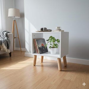 May include: A white, modern side table with four wooden legs. The table has an open shelf with vinyl records, a potted plant, and a tray with headphones. A floor lamp and a couch are in the background.