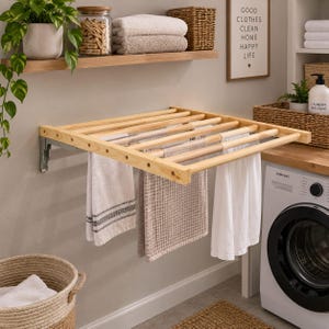 May include: A wall-mounted wooden drying rack with horizontal slats, featuring metal brackets attached to the wall. Three light-coloured towels hang from the wooden rungs. The rack is positioned above a white washing machine in a laundry room.