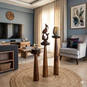 May include: A living room scene featuring wooden sculptures on pedestals, a round jute rug, and a gray armchair. The room has a neutral color palette with a large TV and a framed botanical print. Decorative items include a wooden clock and a vase.