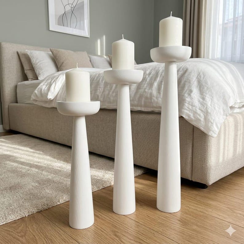 May include: Three white candle holders of varying heights, each with a wide, shallow dish to hold a white pillar candle. The holders have a minimalist design with a conical base and a rounded top. The candles are unlit.