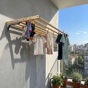 May include: A wall-mounted wooden drying rack with multiple horizontal dowels attached to metal brackets. The rack holds various items of clothing including socks, a t-shirt, a towel, and shirts secured with wooden clothespins on a balcony.