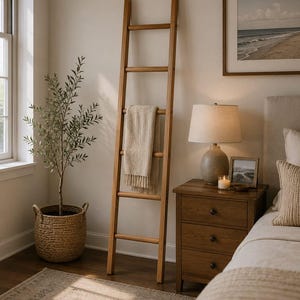 May include: A tall, light brown wooden blanket ladder leaning against a wall. The ladder features five horizontal rungs, with a textured, cream-coloured woven throw blanket draped neatly over the third rung from the top, adding a soft element.