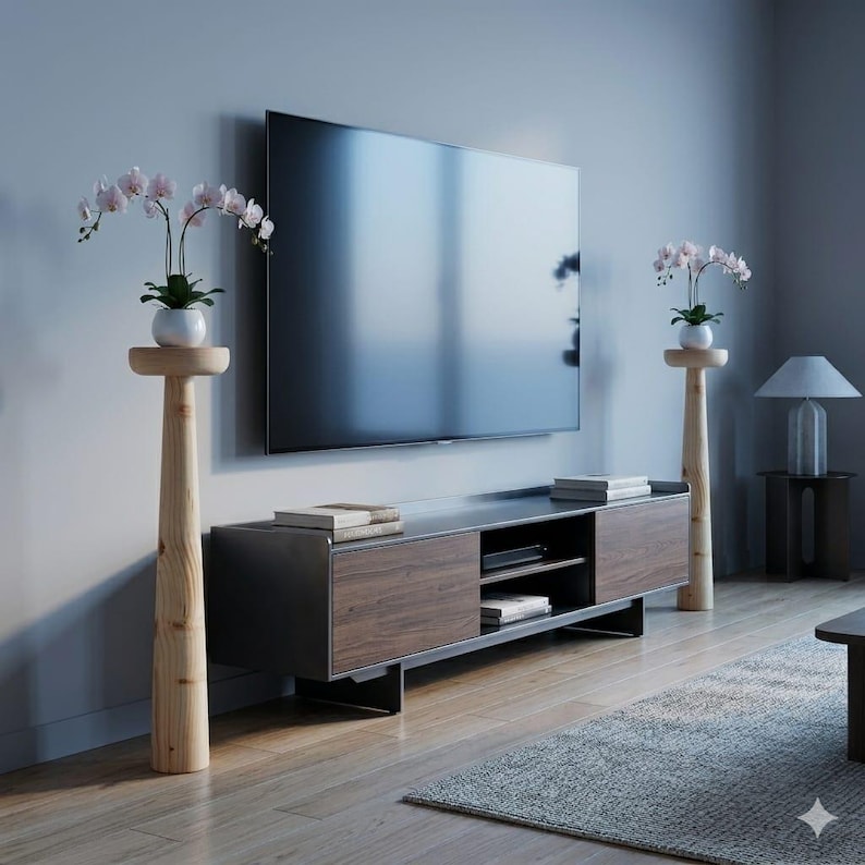 May include: A modern living room setup featuring a large flat-screen TV mounted on a gray wall. A dark wood and metal TV stand sits below. Two wooden plant stands with orchids flank the TV, and a gray rug is on the floor.