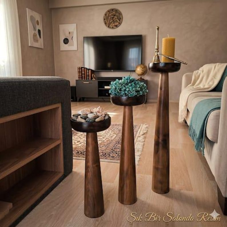 May include: Three wooden pedestal stands of varying heights. The stands have a dark brown finish and a round top. One stand holds a collection of stones and seashells, another has a blue plant, and the tallest holds a candle and a decorative bell.