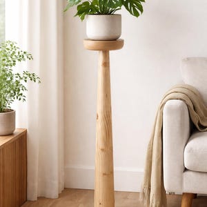 Puede incluir: Un soporte de planta alto de madera clara con una parte superior redonda que sostiene una planta en maceta con hojas verdes grandes. El soporte tiene un diseño delgado y cónico. El fondo incluye una pared blanca, una ventana y un sofá beige.