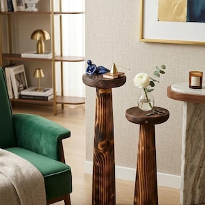 Op de afbeelding: Twee houten bijzettafels met een donkere vlek. Eén tafel heeft een blauw beeldje en een kleine gouden piramide. De andere tafel heeft een vaasje met een witte bloem en een schaar. De tafels staan in een woonkamer.