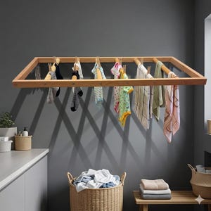 Puede incluir: Un tendedero de madera con ropa colgada. El tendedero está montado en una pared gris y tiene varios artículos de ropa sujetos, incluyendo calcetines, ropa de bebé y toallas. Debajo hay una cesta de mimbre llena de ropa.