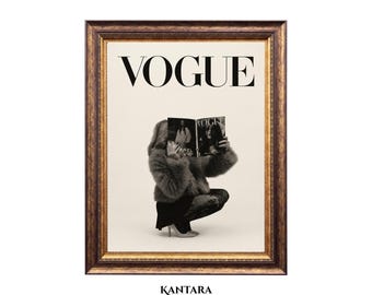 May include: A framed wall art print featuring the word VOGUE in black capital letters at the top. Below, a person in a fuzzy gray jacket and dark skirt crouches while holding an open VOGUE magazine in front of their face. The frame is dark brown and gold.