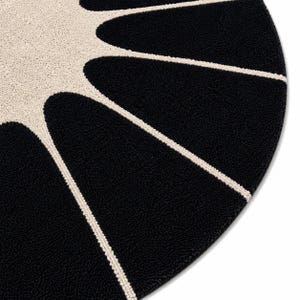 May include: A circular rug featuring a high-contrast design with a central cream-colored sunburst pattern radiating outward into deep black segments. The textured surface displays a dense, tufted pile construction with clean, defined lines.